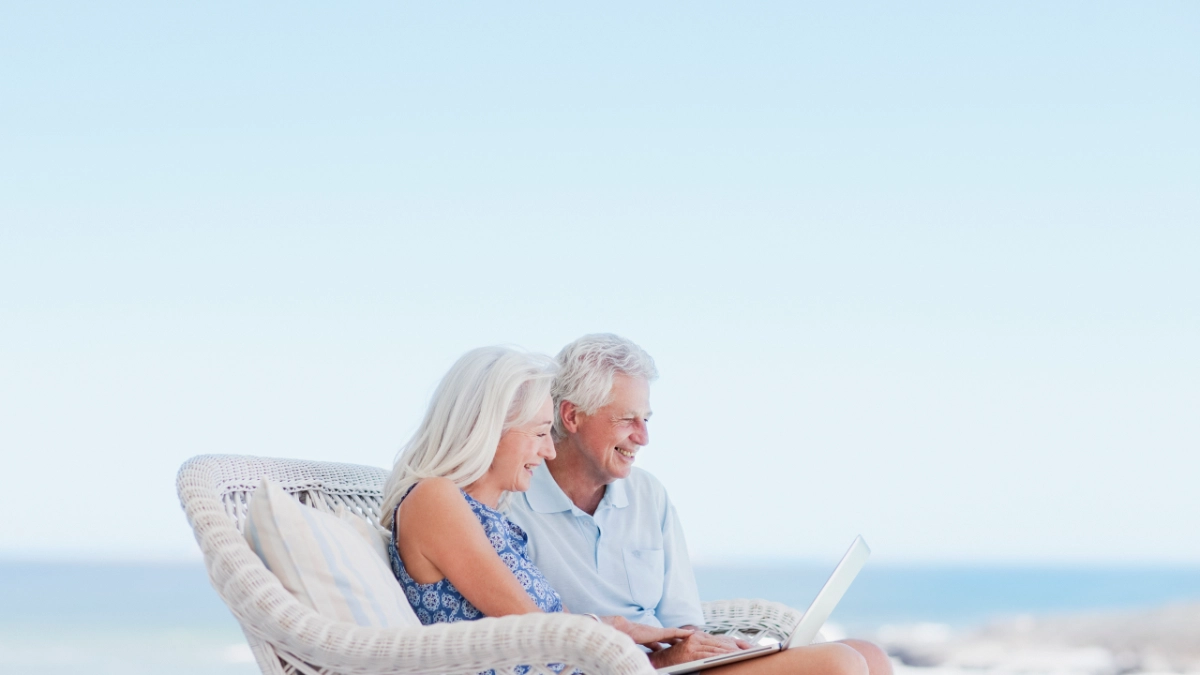 an image of a old couple siiting on a beach checking on their laptop