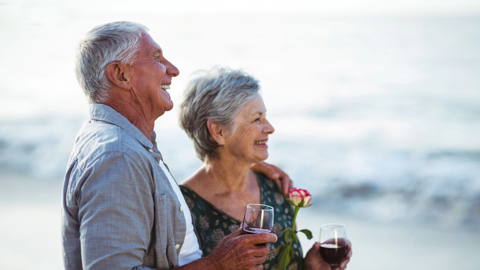 A smiling senior couple standing together on a beach, holding glasses of wine.