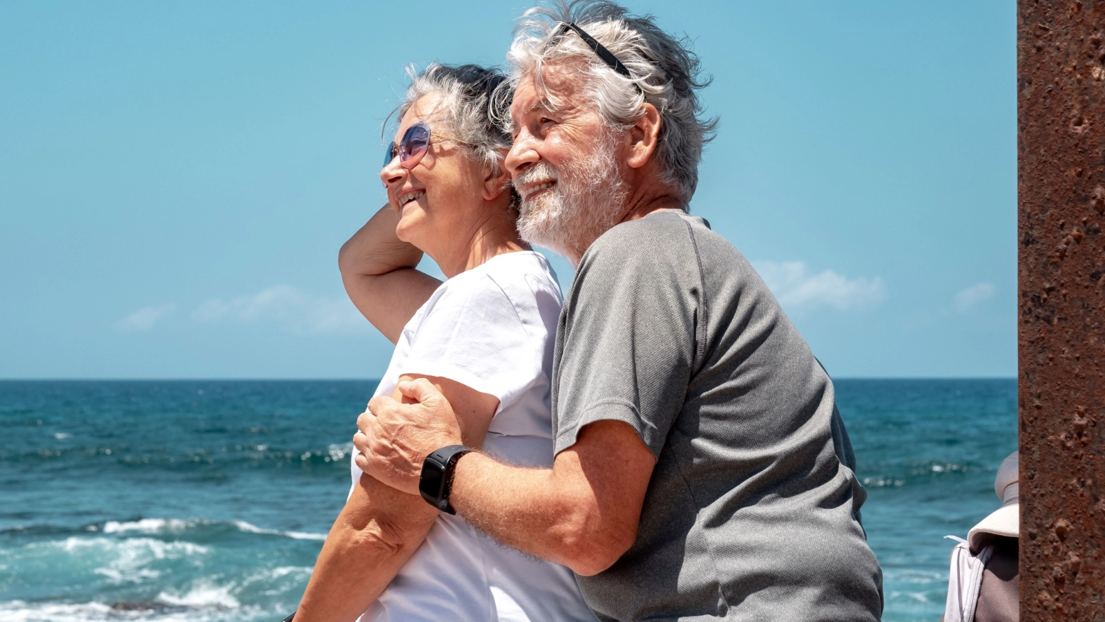 A smiling senior couple sitting together by the ocean on a sunny day.