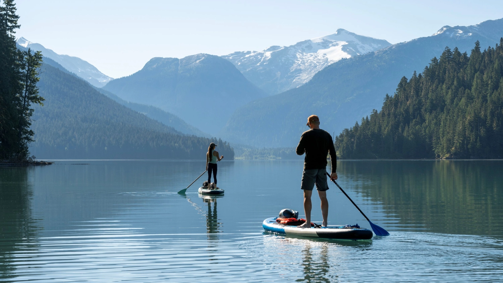 Couple paddle SUP boards across mountain lake, Cheakamus Lake, Whistler.